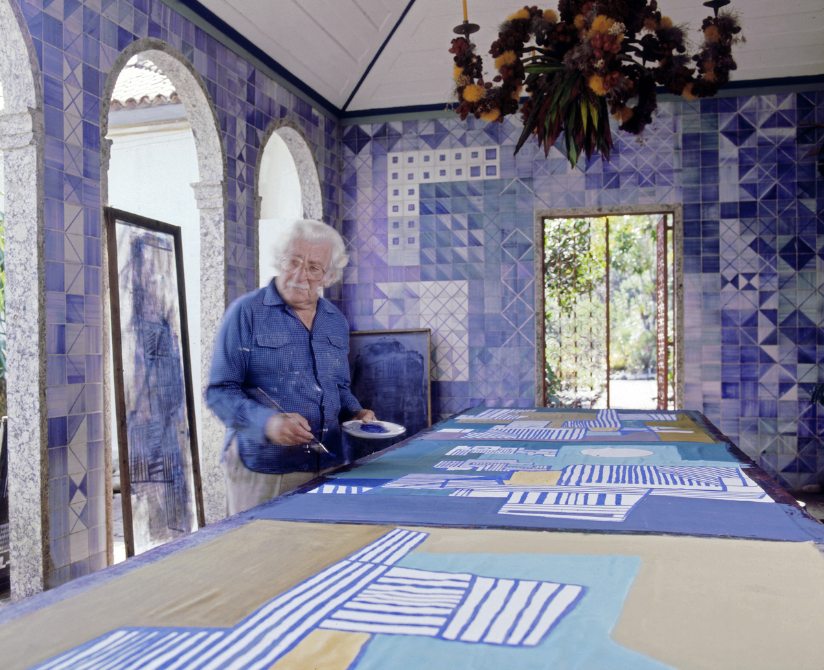 Roberto Burle Marx painting a tablecloth in the loggia of his home, 1980s; the azulejo tile walls and chandelier composed of fruit and flowers on a metal armature are his work. Photograph © Tyba.